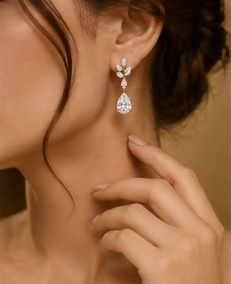 A close-up lifestyle shot of a woman wearing a delicate silver-toned dangle earring with clear gemstones, featuring a small floral cluster and a pear-cut stone. The warm, soft-focus background highlights the jewelry on her ear.3