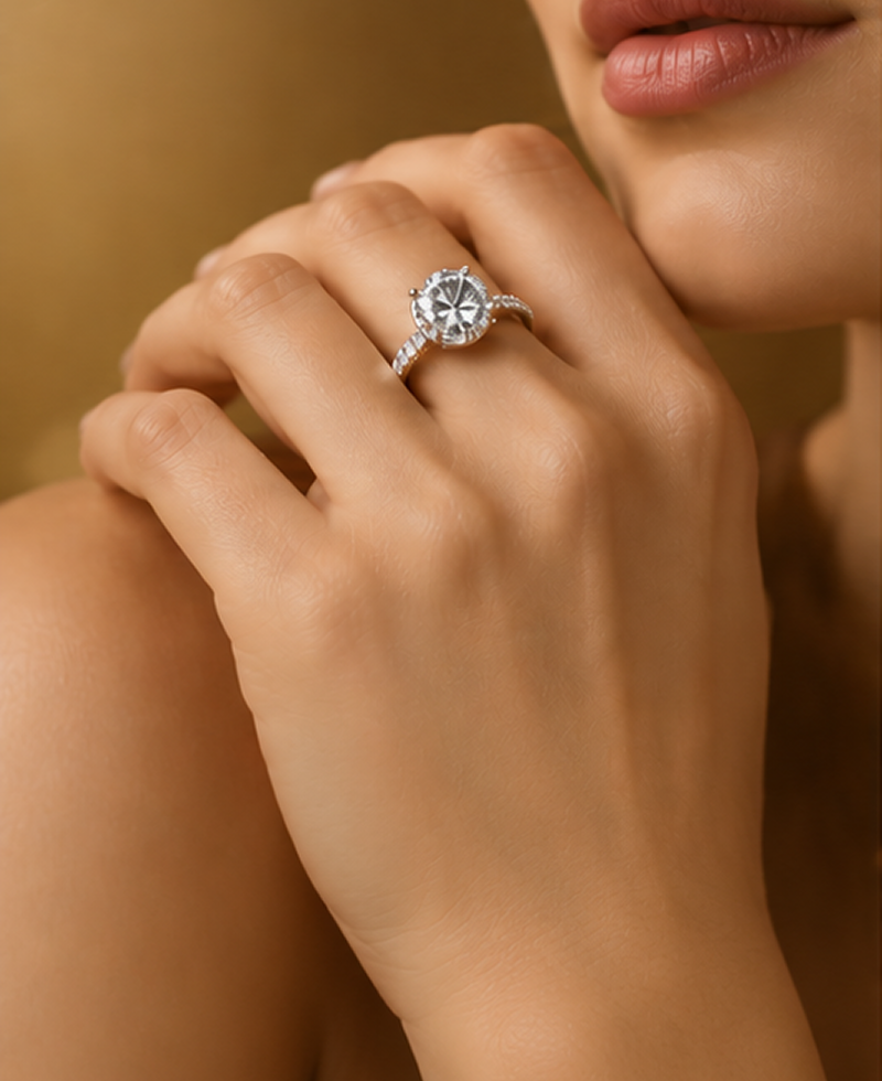 A close-up lifestyle shot of a woman's hand wearing a silver-toned ring with a large round brilliant-cut center stone and smaller surrounding stones, set against a warm, soft-focus background.4