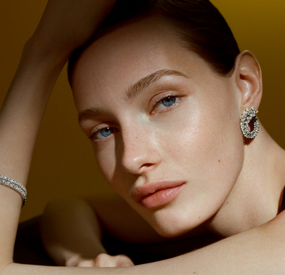 A close-up portrait of a woman with blue eyes wearing a silver crescent-shaped hoop earring with black and white stones, and a matching silver bracelet on her wrist, set against a warm golden-brown background.