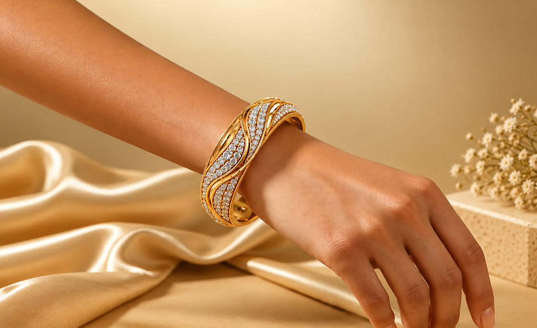 A close-up of a person's hand and wrist wearing a wide gold bangle bracelet paved with small clear stones, posed over draped champagne satin fabric with a small floral arrangement in the background.