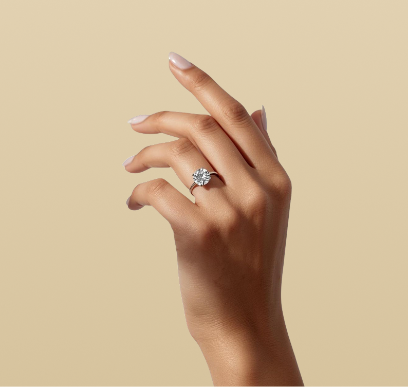 A close-up of a hand with manicured nails wearing a silver-toned halo ring with a round clear gemstone, set against a solid beige background.